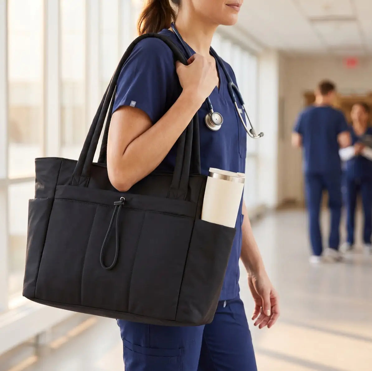A nurse in navy scrubs carrying the Everyday Laptop Tote with an insulated tumbler in the side pocket